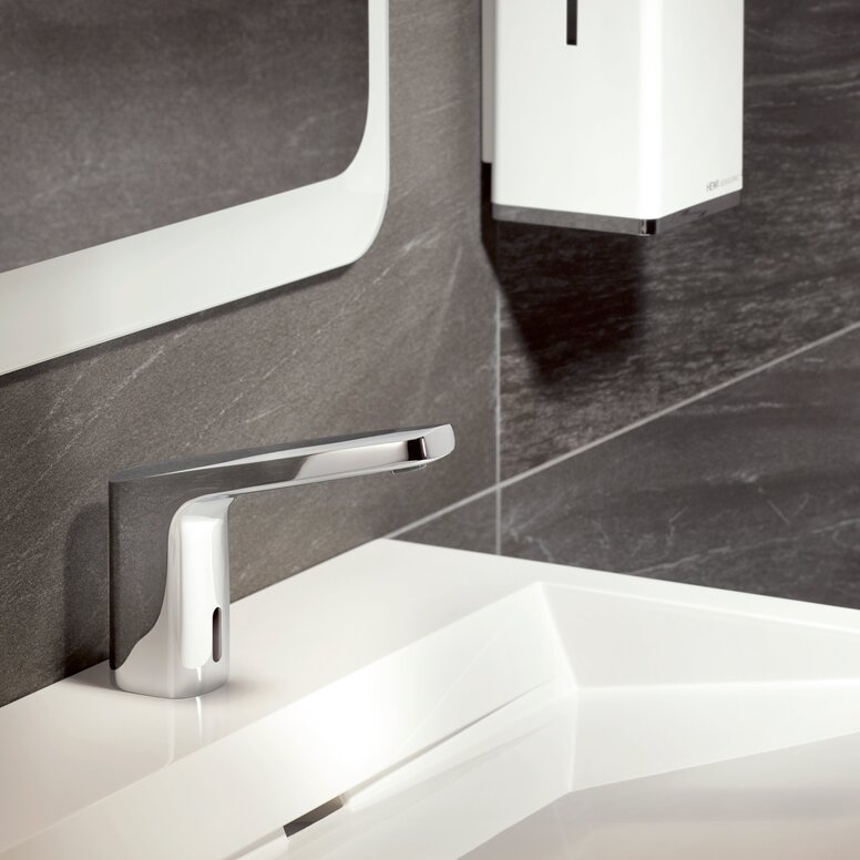 HEWI Fittings AQ SENSORIC Electronic Washbasin Fitting A chrome-plated touchless tap on a white washbasin against a backdrop of dark grey stone tiles