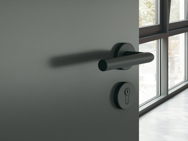 HEWI System 162 Türdrücker Dunkelgraue pulverbeschichtete Türklinke mit runder Rosette an dunkelgrauer Tür, dahinter ein Arbeitszimmer mit Schreibtisch und Stuhl