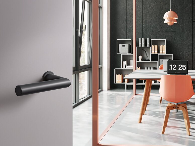 HEWI System 162 lever handle A black synthetic material door handle on a grey door; behind it, an office with a desk and chair in front of a dark grey wall