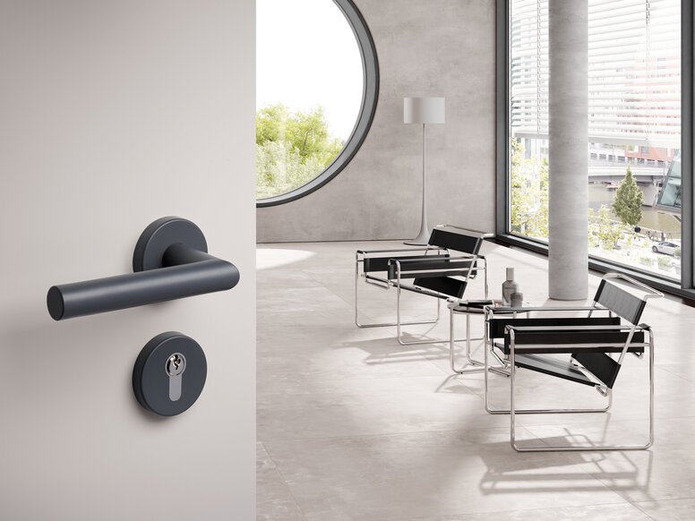 HEWI System 162 lever handle Dark grey lever handle with a round rose and matching keyhole escutcheon on a light grey door, with black armchairs and large windows in the bright room beyond