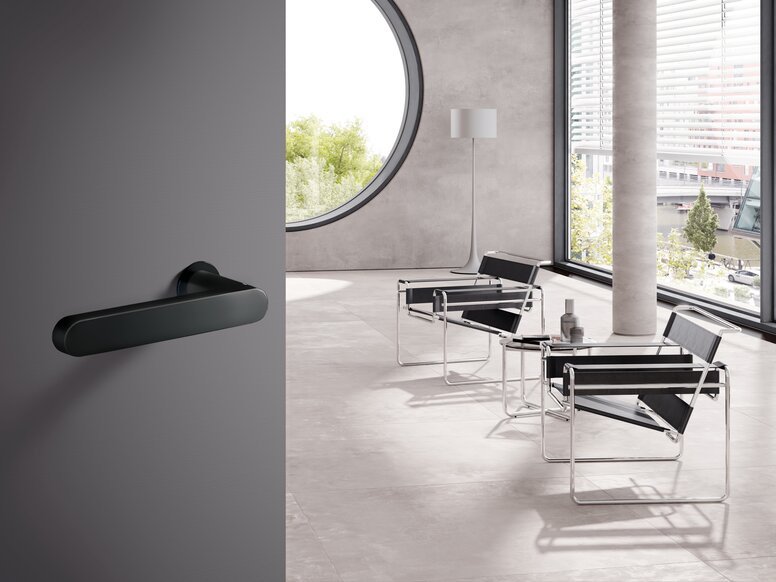 A black powder-coated door handle fitted to a grey door; behind it, a bright room with two chairs
