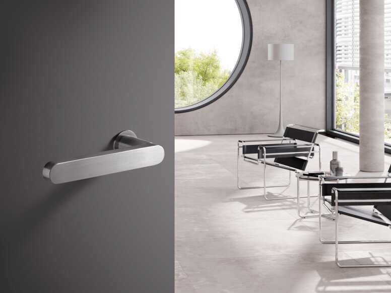 HEWI Range 270 lever handles A stainless steel door handle on a grey door; behind it, a bright room with chairs