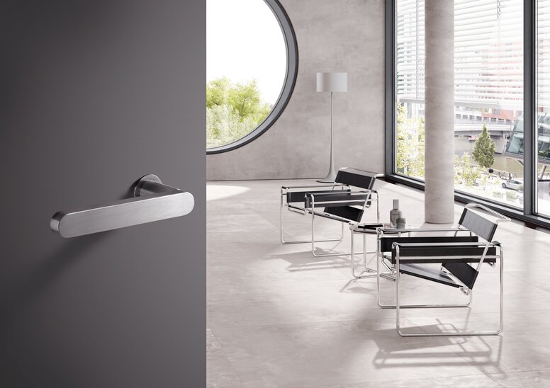 A stainless steel door handle on a grey door; behind it, a bright room with chairs