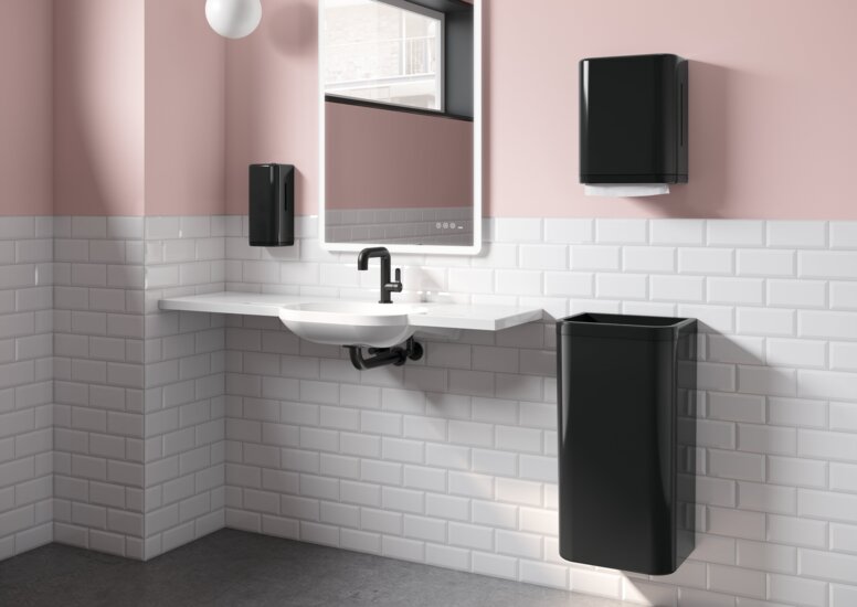 A white washbasin with a black paper towel dispenser and wastebin next to it, in a bathroom with white tiles and pink walls