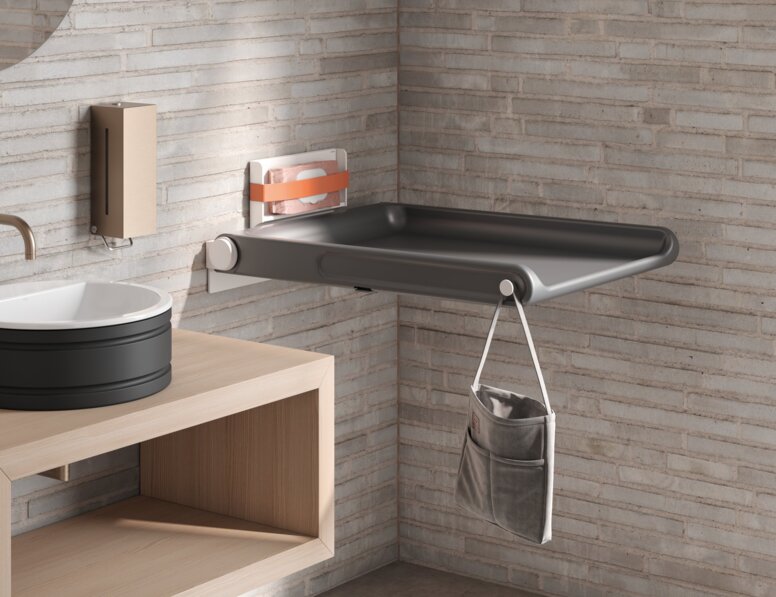 HEWI Serie 480 Babywickeltisch Klappbarer Babywickeltisch mit Seitentasche zur Aufbewahrung und einem Haken an heller Ziegelwand montiert
