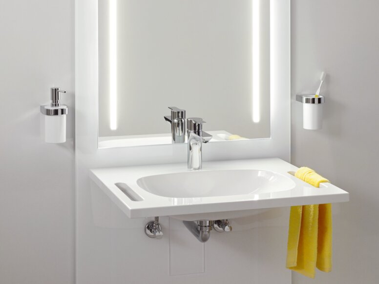 A white washbasin with a tap, a recessed handle on the side and an illuminated mirror with two light strips on a light grey wall