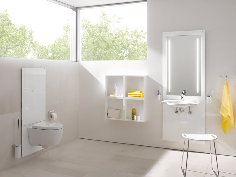 A white WC module in a bright bathroom with an illuminated mirror above the washbasin, in front of which stands a stool with a white seat