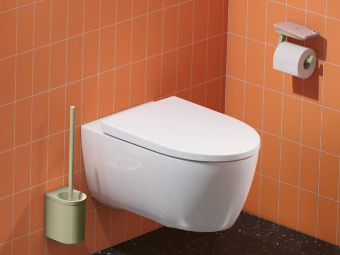 Washbasin with green fittings, a round mirror and cosmetic mirror, beside a wall-hung toilet with a toilet roll holder against green and orange tiles