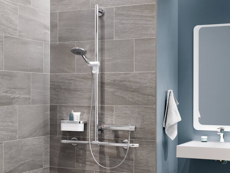 A hinged shower seat with a support rail, a thermostatic mixer and a shower head against a grey tiled wall, next to which is a washbasin area against a blue wall