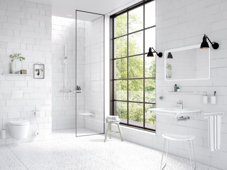 HEWI System 162 Bathroom Interior Bright bathroom with white wall tiles, a level-access shower behind a glass partition, a wall-hung toilet and a white washbasin beneath a mirror, complemented by white grab rails and a large black-framed grid window